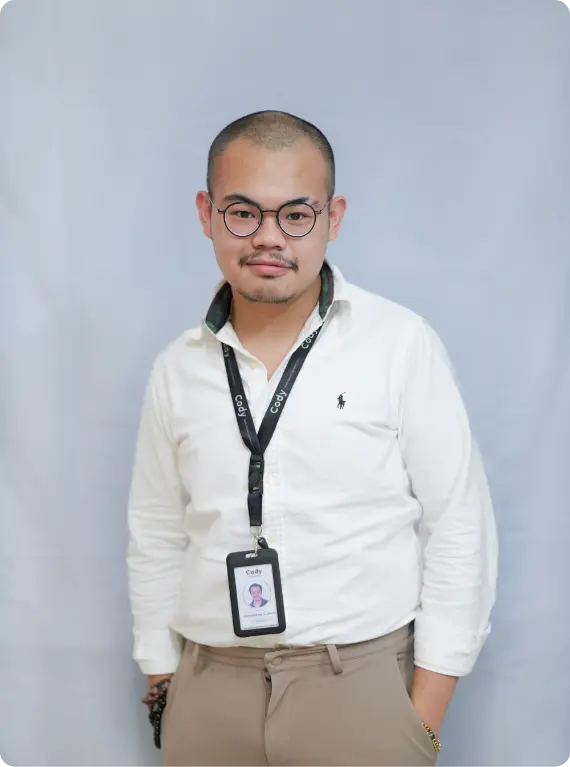 Portrait of Garfield Lim, Technical Director, standing with hands in pockets, wearing glasses, a white long-sleeve shirt, and a black lanyard with an ID, against a plain gray background.
