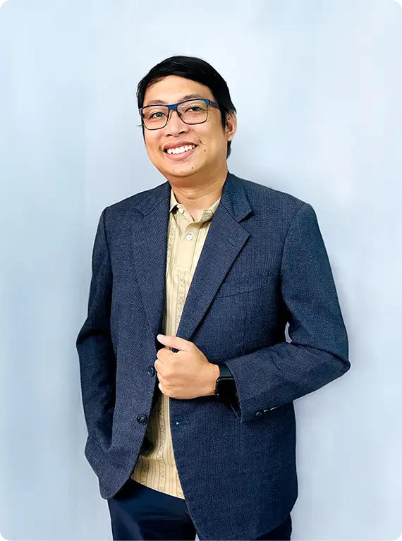 Portrait of Christian Ivan Igot, Technical Director, standing with hands in pockets, wearing glasses, a white long-sleeve shirt, and a black lanyard with an ID, against a plain gray background.